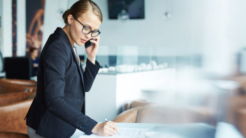 Blond confident businesswoman going over details of contract talking with partner on phone during her coffee break in modern light lounge area